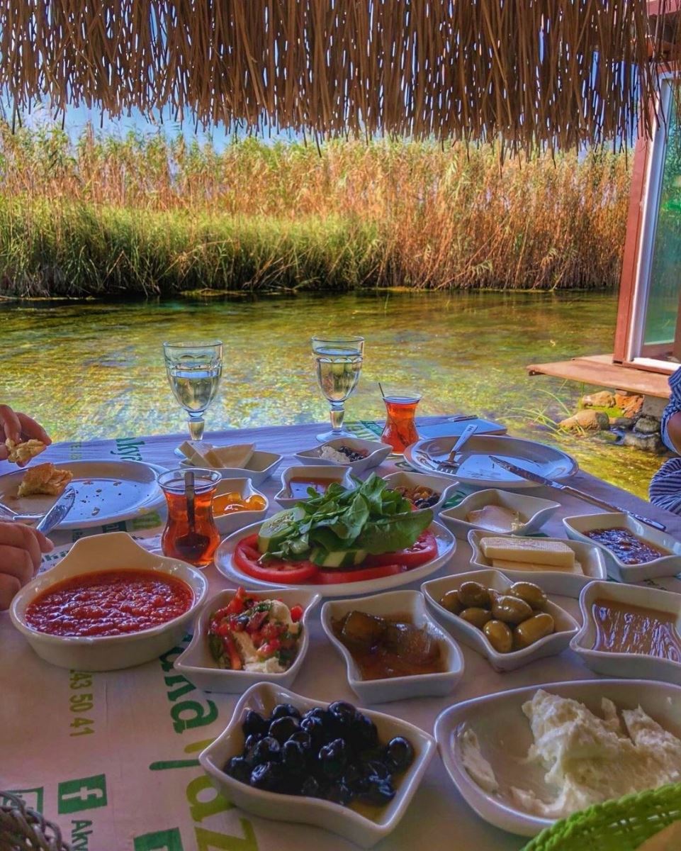 Azmak Deresi kenarındaki küçük restoranın terası, önde zeytinyağlı meze tabakları, arka planda dere ve kamışlıklar