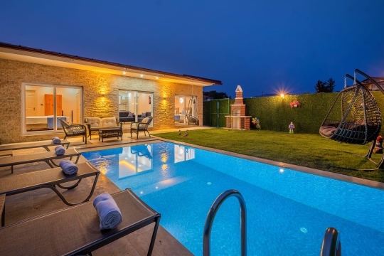 Fethiye Villa Vimo’nun gece aydınlatmalı özel yüzme havuzu, taş dış cephesi ve havuz başı dinlenme alanı.