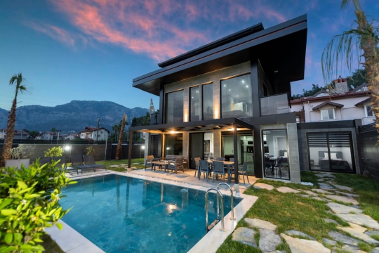 Yemyeşil çim bahçe, taş yürüyüş yolu ve arka planda çam ormanlarıyla çevrili villa peyzaj alanı.