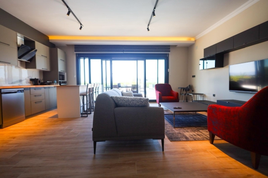 Lüks gri köşe koltuk, kırmızı berjer, modern aydınlatmalar ve bahçeye açılan cam kapılı salon