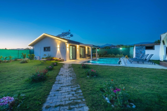 Yemyeşil çim bahçede kilit taşlı yürüyüş yolu, akşam aydınlatmalı yüzme havuzu ve villa mimarisi.