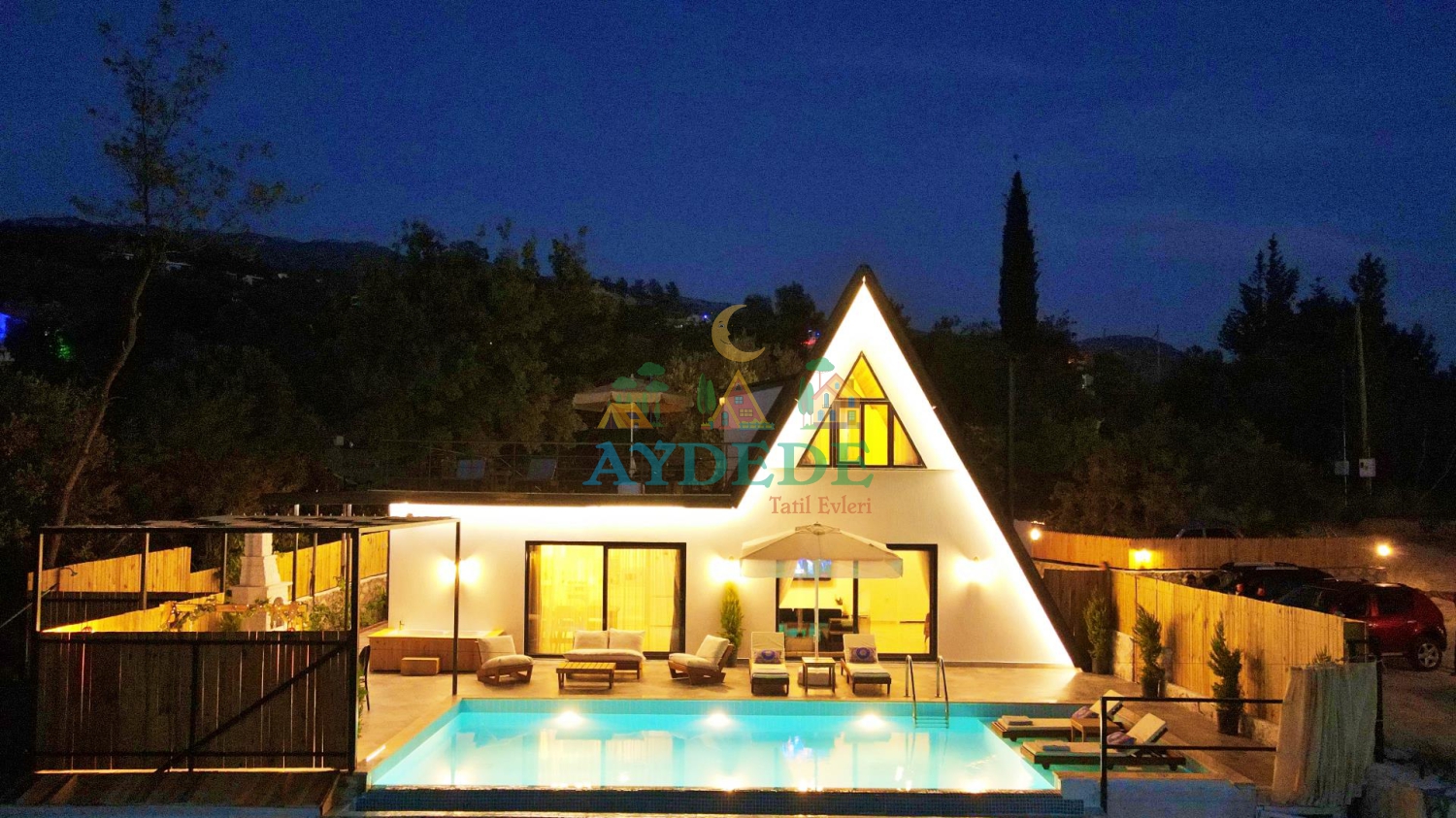 Gece aydınlatmaları altında ışıldayan özel yüzme havuzu, ahşap şezlonglar ve modern üçgen A-frame mimarili villa dış cephesi.